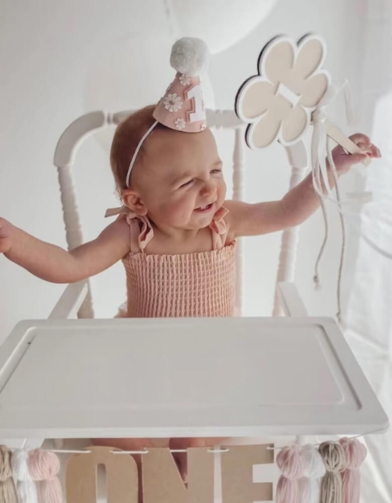 A happy baby girl in an adorable pink onesie wearing Momo Party's Daisy Applique felt birthday hat on her head. In one of her hands, she is holding a flower shaped wooden wand with number 1 on it. She's happily waving the want sitting on a white vintage inspired highchair which is adorn with ONE wool tinsel highchair garland celebrating her first birthday.