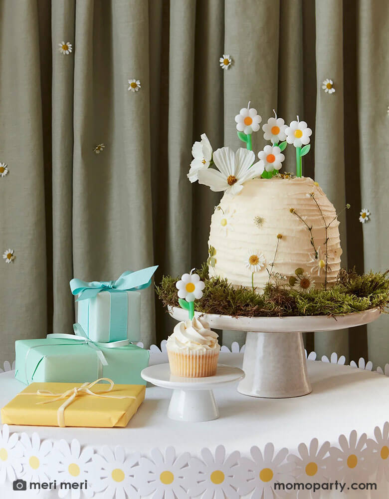 A wild daisy themed birthday party table featuring a buttercream cake decorated with real daisies on the side and topped with Momo Party's daisy candles from Meri Meri. There's also a daisy paper decoration wrap around the table which has some wrapped gifts on it. In the back is a curtain decorated with real daisies flowers, making this an adorable inspo for girl's daisy or garden themed birthday party.