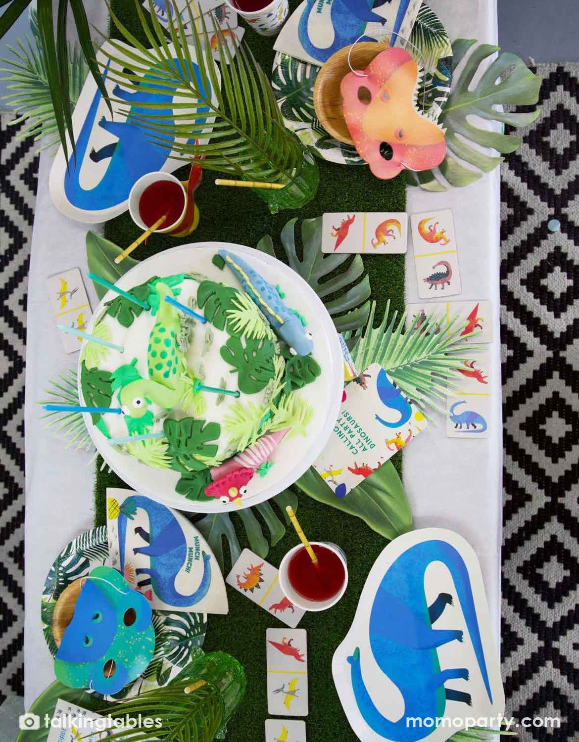 A dinosaur themed kids party table setup featuring multiple dinosaur themed party supplies from Momo Party including the artificial green grass table runner, dinosaur shaped paper plates, napkins, party cups with red drinks, temporary tattoos, and masks. The table is decorated with green palm leaves scattered around the table setting.
