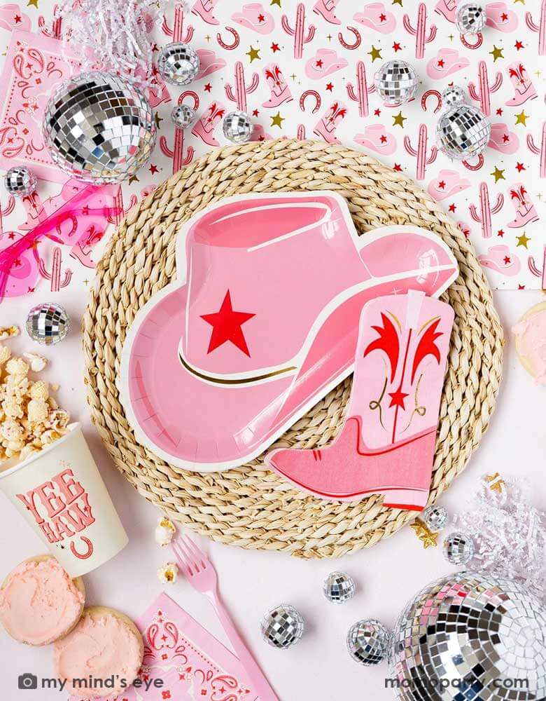 A cowgirl rodeo Western themed party table features pink cowgirl themed party supplies from Momo Party including pink cowgirl boot shaped napkins, cowgirl hat shaped plates, pink bandana small napkins, yeehaw party cups and cowgirl pattern paper table runner, along with festive disco ball decorations, heart shaped sunglasses and treats, this makes a perfect inspo for kid's cowgirl themed birthday party or a bachelorette party celebration!