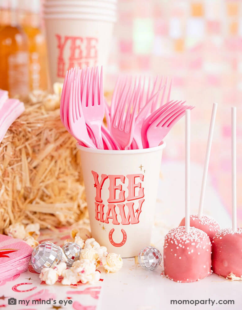 A cowgirl rodeo Western themed party table features pink cowgirl themed party supplies from Momo Party including 12oz pink YEEHAW party cups with pink utensils, cowgirl boot shaped napkins on the cowgirl pattern paper table runner, along with hay stacks, some small disco ball decorations and pink cake pops and popcorn, making this a perfect inspo for kid's cowgirl themed birthday party or a disco cowgirl bachelorette party celebration!