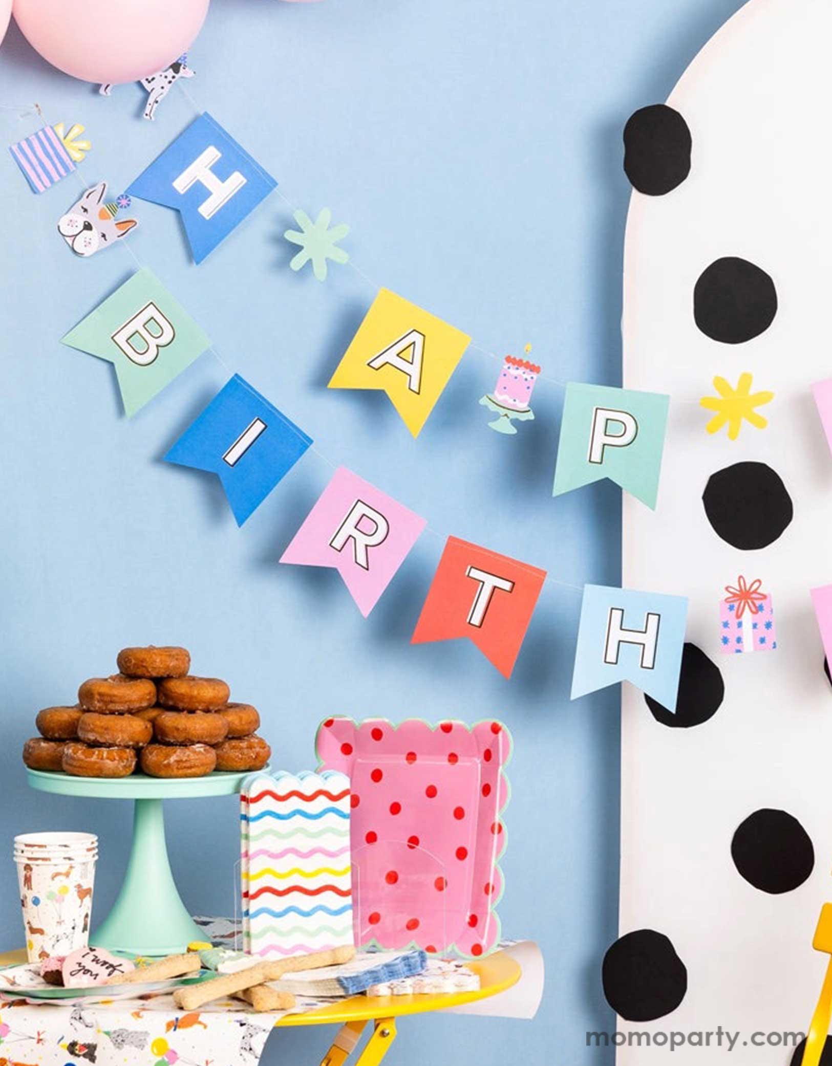 A corner of a festive and cheerful dog themed party featuring Momo Party's dog party tableware collection including dog themed party tablewares, the dog party Happy Birthday banner hung over a polka dotted party backdrop and party balloons. On the table there are some sugar cookies, treats and a donut tower, ready for a fun and playful birthday celebration for a pet lover!