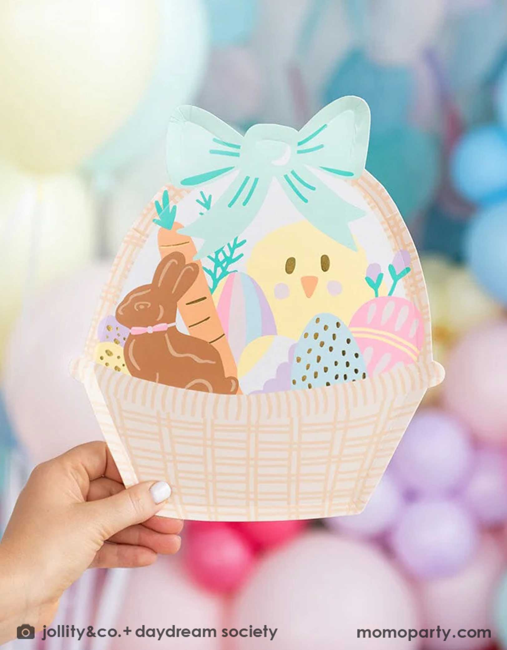 A little kid's hand holding Momo Party's Easter Fun basket shaped plate by Daydream Society. In the back there's a wall of balloons in pastel spring inspired colors, making this an adorable Easter party decoration.
