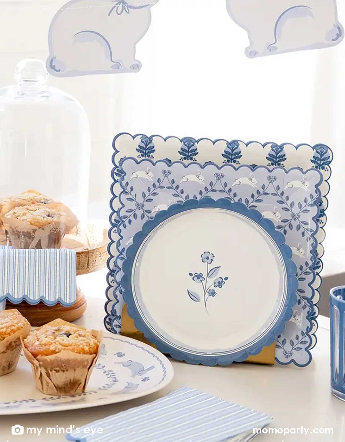 An elegant Easter brunch table setting featuring multiple blue bunny themed tablewares from Momo Party including the blue oval bunnies bamboo serving tray with muffins on it, blue bunny shaped napkins, blue striped guest napkins, bunnies pattern square plates and blue floral scallop round plate. With a blue bunny shaped banner hung above, this makes a great inspo for a beautiful Easter table setting idea.