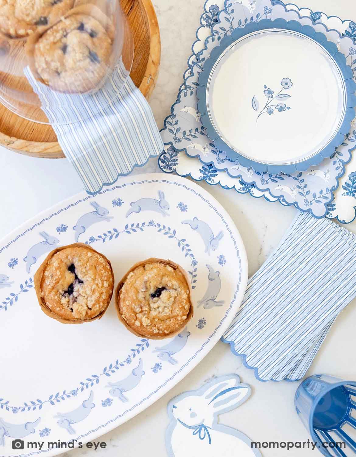 An overhead shot of an elegant Easter brunch table setting featuring multiple blue bunny themed tablewares from Momo Party including the blue oval bunnies bamboo serving tray with muffins on it, blue bunny shaped napkins, blue striped guest napkins, bunnies pattern square plates and blue floral scallop round plate.