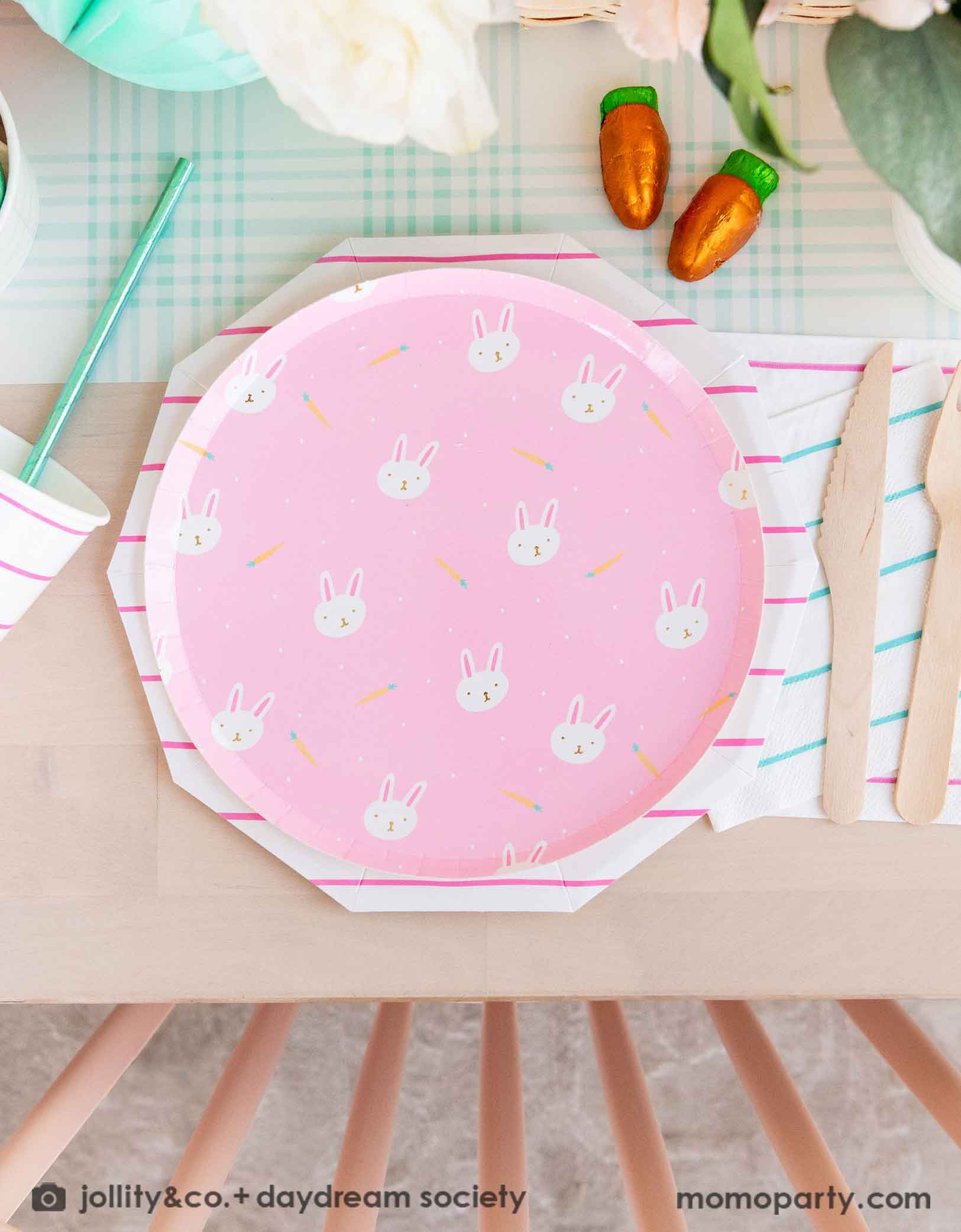 A beautiful Easter brunch table features Momo Party's pink Easter fun small plates along with striped party plates, cups, and napkins. In the middle of the table lays a blue gingham table runner which has baskets of spring flower arrangement and carrot shaped candies, making this an adorable inspo for a kid's friendly Easter party table setting.