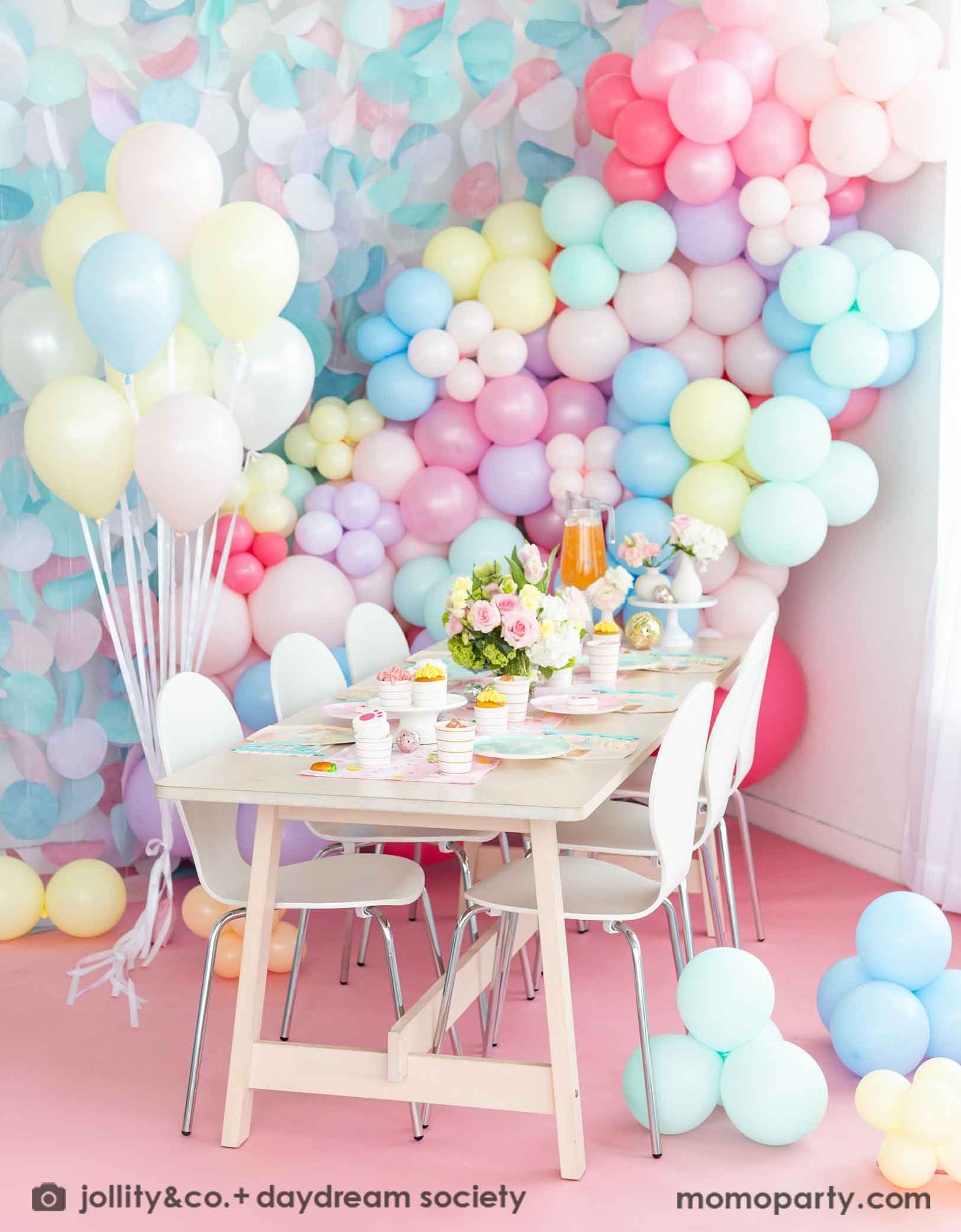 A festive Easter party table features Momo Party's Easter fun small plates along with striped party plates, cups, and napkins. In the back of the table there's a pastel balloon wall with pastel paper decorations, making this an adorable inspo for a Easter party decoration set up.