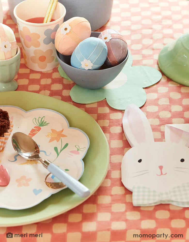 An Easter party table with Easter eggs adorned with Meri Meri's Easter themed tattoo stickers, on the table are Momo Party's Easter themed plates and napkins in soft pastel colors including bunny shaped napkins which are coordinated with the charming daisy shaped napkins, makes this an adorable tablescape idea for a spring gathering.