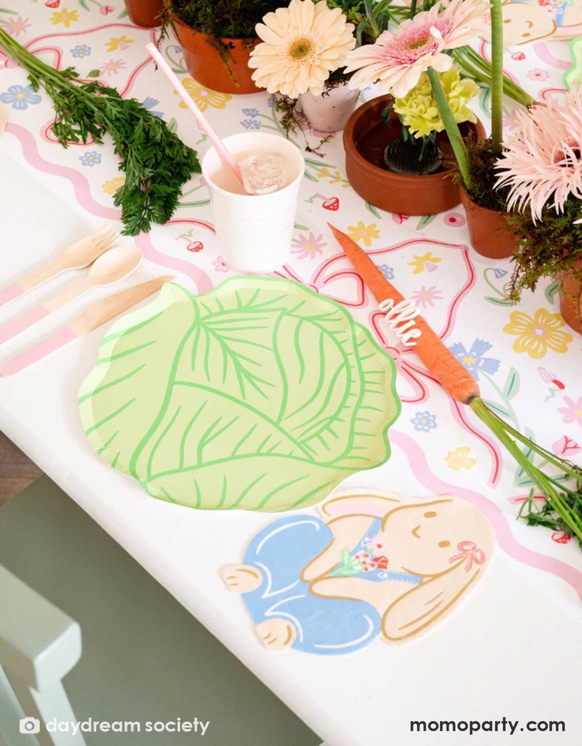 A spring inspired party table setting featuring colorful Easter themed tablewares from Momo Party including cabbage shaped plates, Easter bunny shaped napkins, matching party cups and utensils over a floral tablecloth with potted plants in the middle as centerpiece.