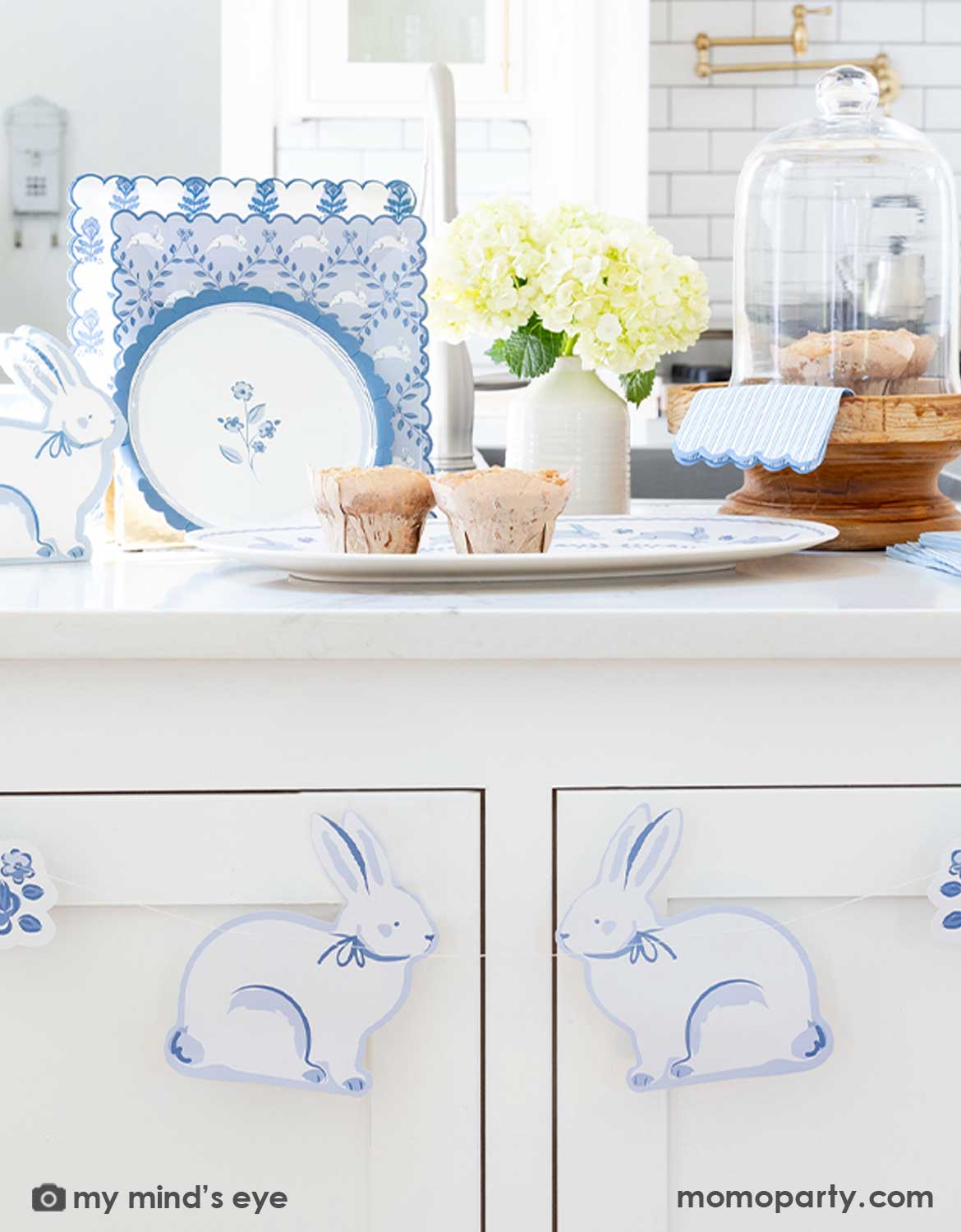 An elegant Easter brunch table setting on a white kitchen counter featuring multiple blue bunny themed tablewares from Momo Party including the blue oval bunnies bamboo serving tray with muffins on it, blue bunny shaped napkins, blue striped guest napkins, bunnies pattern square plates and blue floral scallop round plate. With a blue bunny shaped banner hung underneath, this makes a great inspo for a beautiful Easter table setting idea.