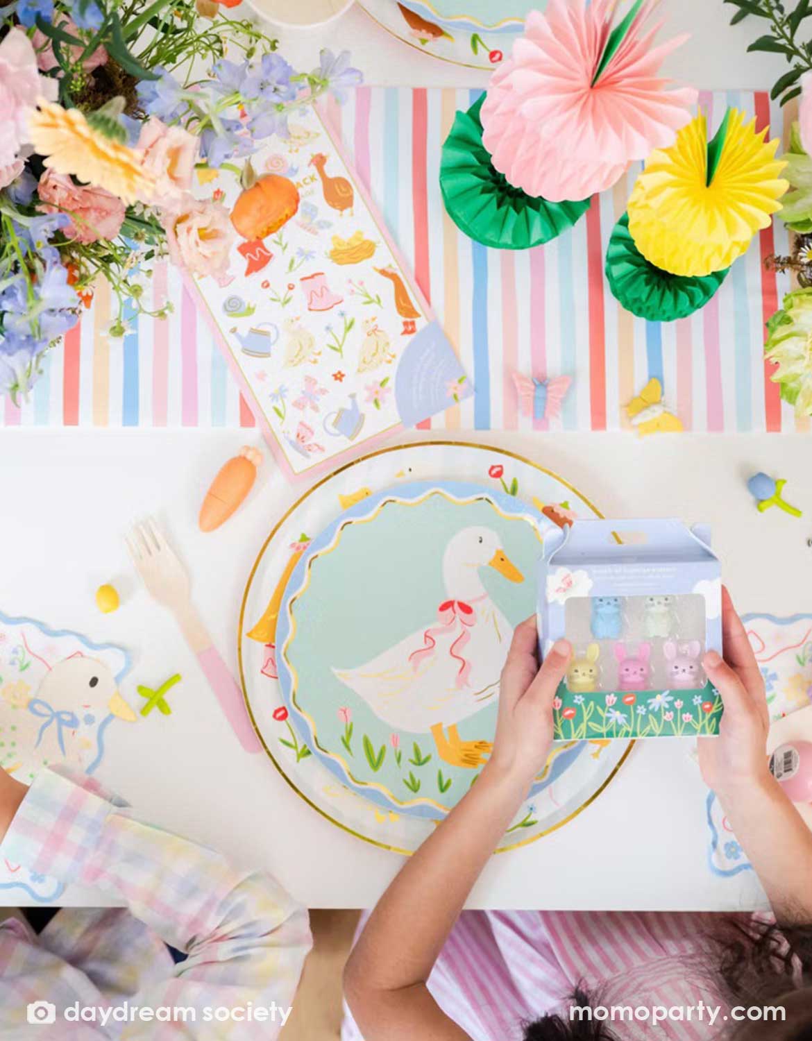 An overhead shot of a children's Easter table setting with colorful flowers, tulip honeycomb decorations, goose themed party plates, napkins, party cups, pink cutlery and multiple kid's Easter small gifts with bunny designs including mini erasers, sticker sheets, carrot shaped mini pens on a colorful striped runner. A perfect idea for an Easter celebration, a baby shower, kid's Silly Goose themed birthday party or a spring gathering.