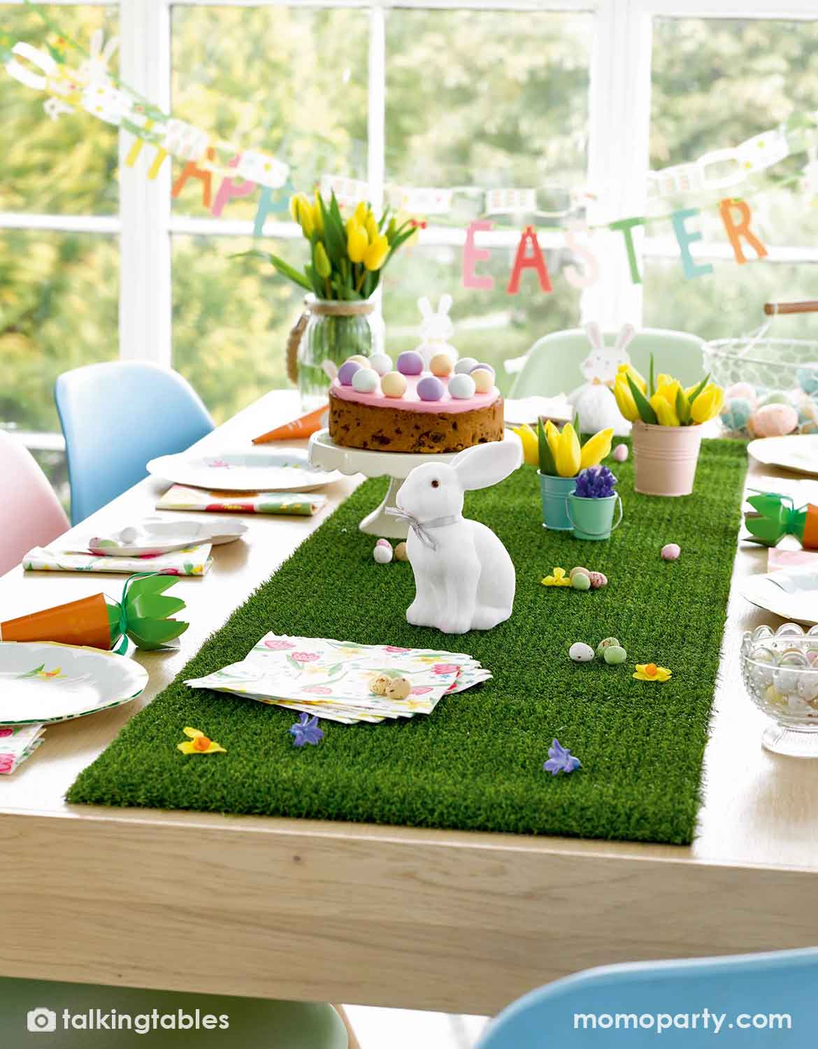 A styled Easter dining table featuring Momo Party's artificial green grass table runner runner, a white ceramic bunny figurine, pastel eggs, yellow tulips, and a small cake, set in a bright room with a window and 'Happy Easter' banner in the background.