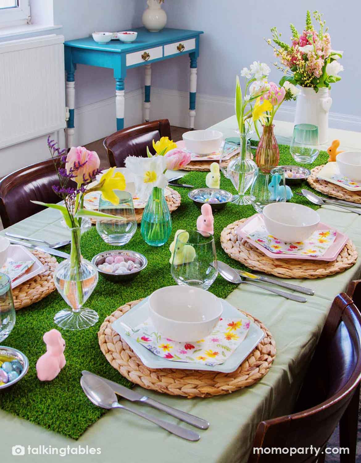 A styled dining table set for a spring or Easter meal, featuring multiple party supplies from Momo Party including green artificial grass runner, woven placemats, white ceramic bowls, pastel paper plates, floral napkins, and small bunny decorations in pastel colors, with flowers and glassware in the background.