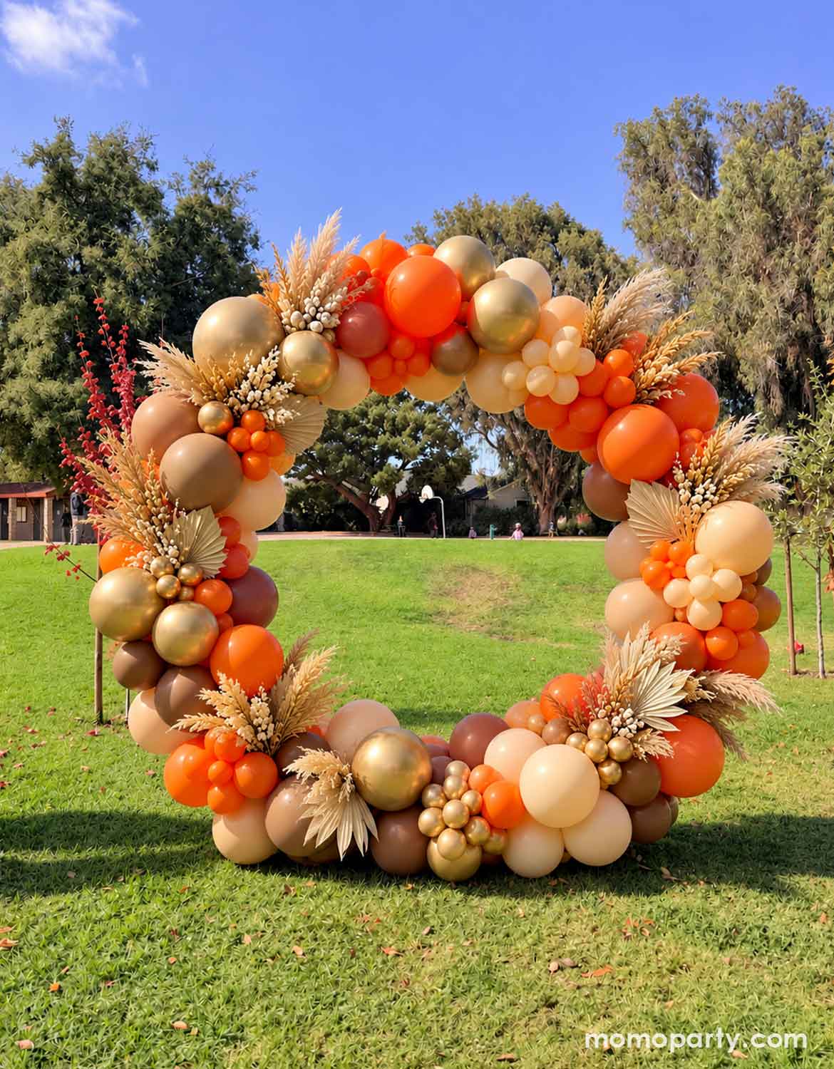 An outdoor balloon arch decoration by Momo Party made of orange, peach, beige, and metallic gold balloons, styled with dried grasses and small floral accents on a green lawn under a blue sky. Perfect for a fall harvest themed birthday party, a Thanksgiving celebration or a boho rustic wedding in autumn.