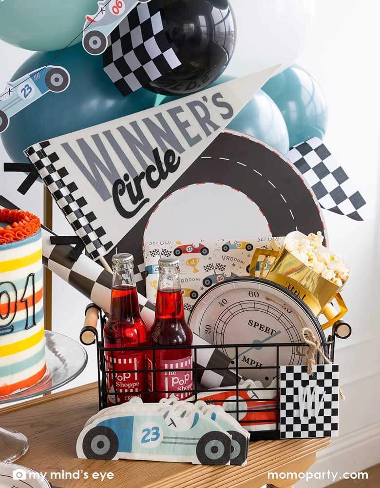 A party occasion bin filled with race car themed party supplies from Momo Party including race track placemats, speedometer shaped paper plates, checkered table runner, race car pattern square plates, race car shaped napkins, and a felt "Winner's Circle" party pennant. Around the basket is a colorful buttercream cake and some party balloons and checkered flags decorated behind the party table. Making this a perfect inspo for kid's race car themed celebrations.