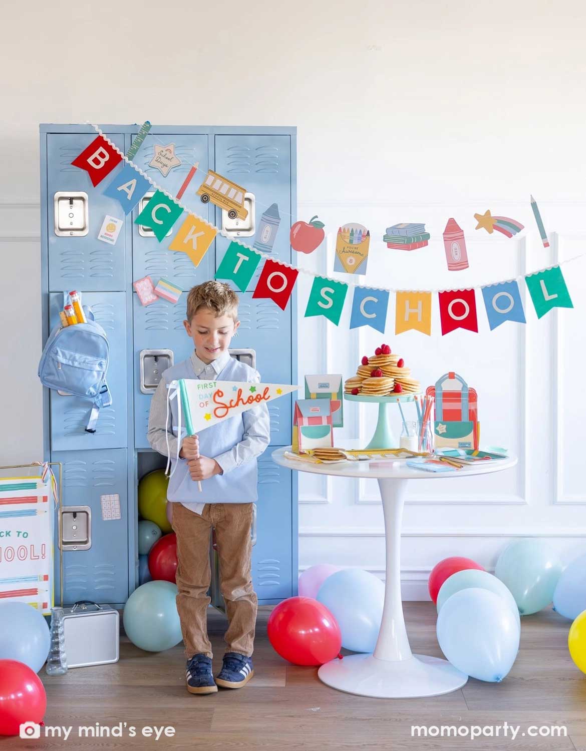 A boy holding the first day of school party pennant in a back to school party set up featuring various school themed party supplies and decors from Momo Party including school themed plates, treat boxes, party pennant, back to school felt garland along with school icons party banner hung above the light blue locker selves. A party table with pancakes on a cake stand next to some colorful backpack shaped treat bags. Along with colorful balloons spread across the room.