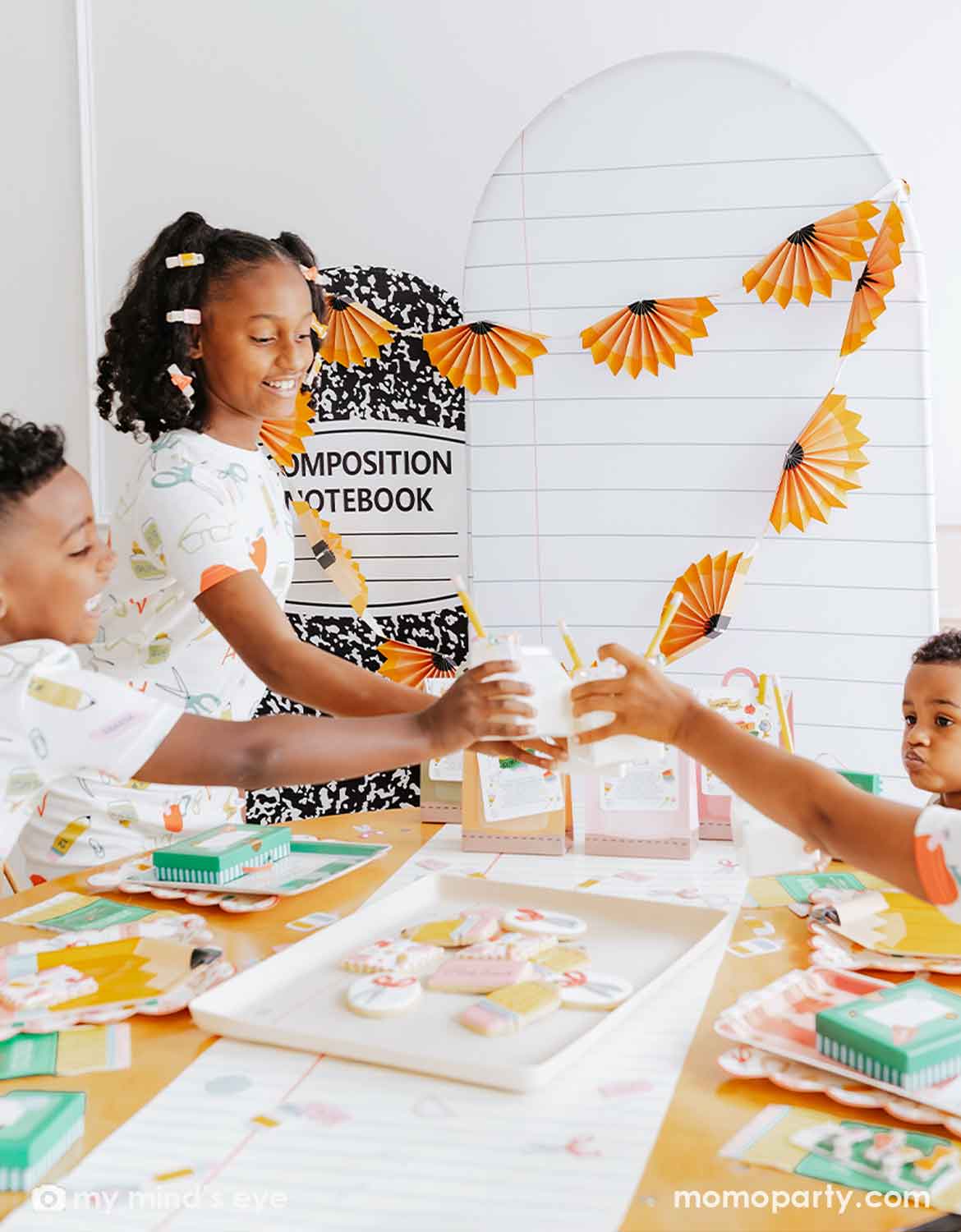 Three children in their school themed pajamas gathered around a table at a first day of school celebration party, cheering with each other with clear milk box shaped cups. The scene is decorated with Momo Party's pencil paper fan banners, a white arched backdrop with school notebook lines design, and a black-and-white patterned composition notebook on the wall behind them. On the table, there are multiple school themed party tablewares including notebook shaped plates, notebook table runner, and more.