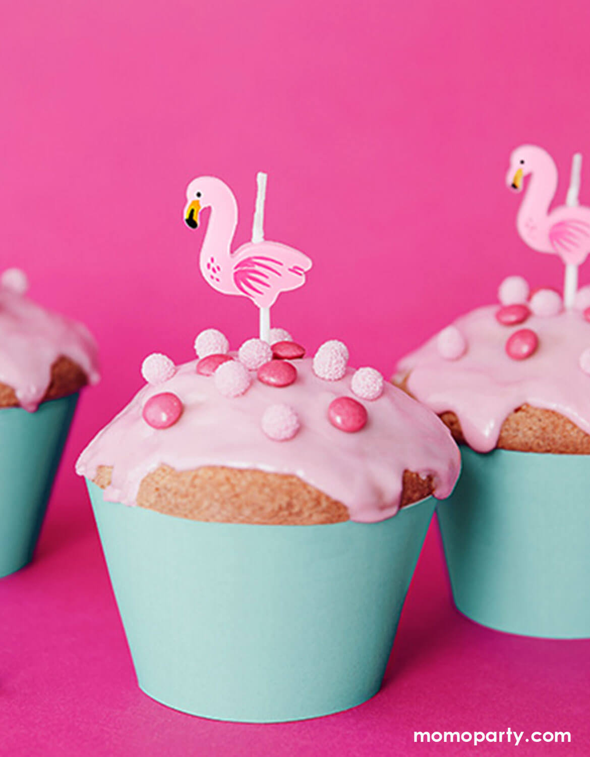 Some cupcakes with pink frosting, and white polka dot chocolates wrapped in mint cupcake wrappers, topped with Momo Party's flamingo shaped candles on a pink background.