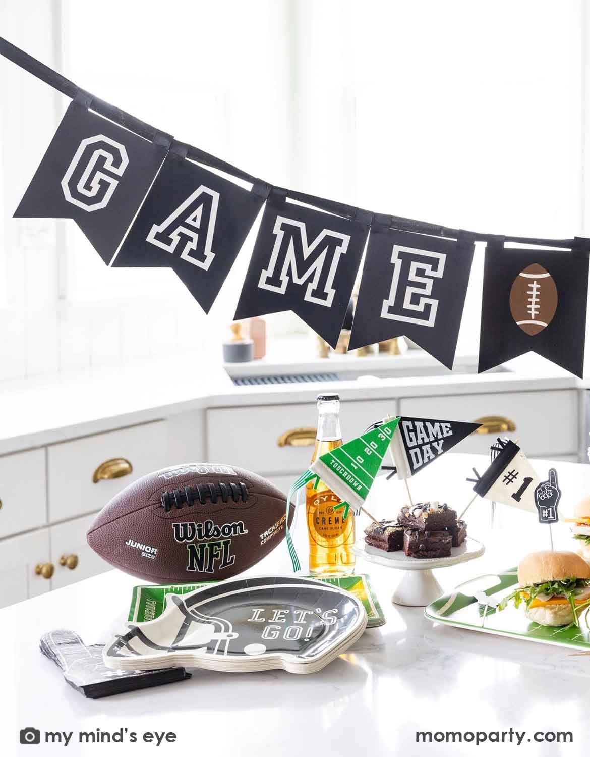 A football-themed party setup with a black banner reading 'GAME Day' and a football icon, a brown Wilson NFL football, a silver tray with a football silhouette, a green and white 'Game Day' cup, and food on a white table in a bright kitchen setting.