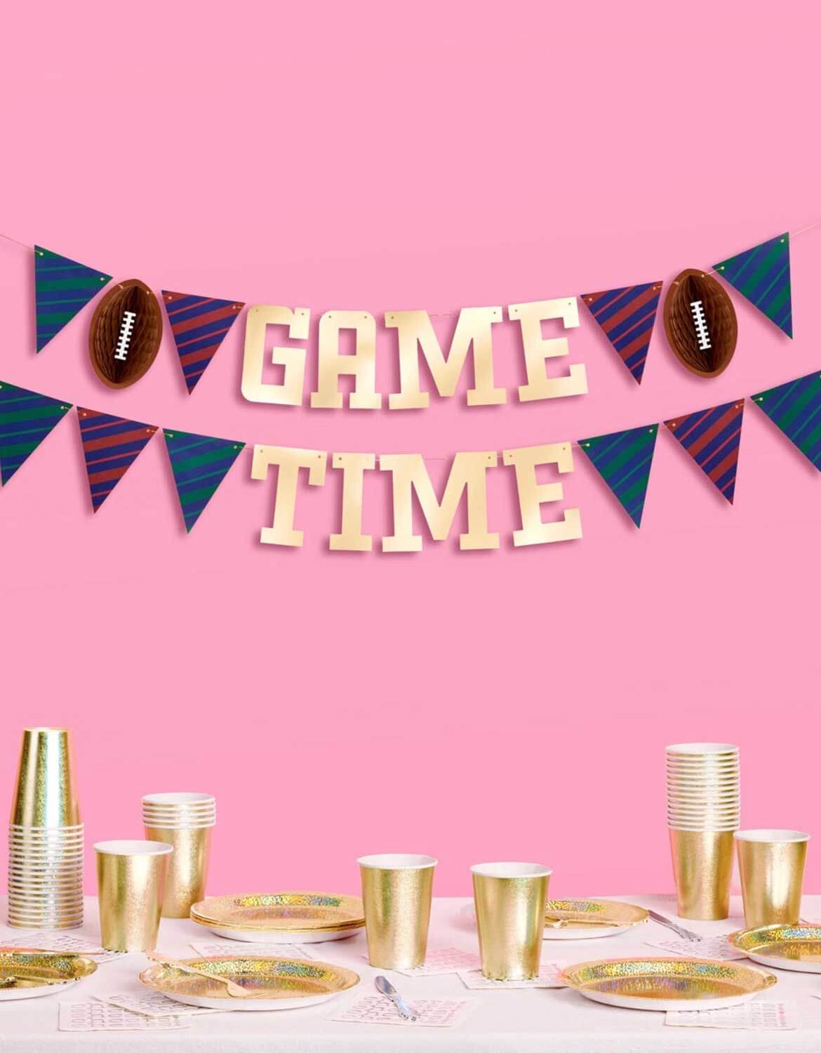 Momo Party's 4.5' football gold foil game time banner hung above a table full of gold themed tablewares, against a pink background.
