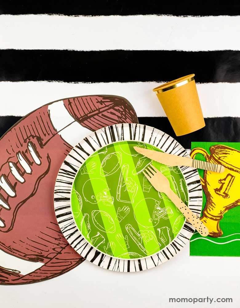 A modern yet festive football party tablescape featuring football themed tableware from Momo Party including the football shaped paper placemat, the touchdown dessert plates, the trophy shaped napkins on a black striped table runner, making this a perfect inpo for a kid's football themed birthday party or a Superbowl party table decoration.