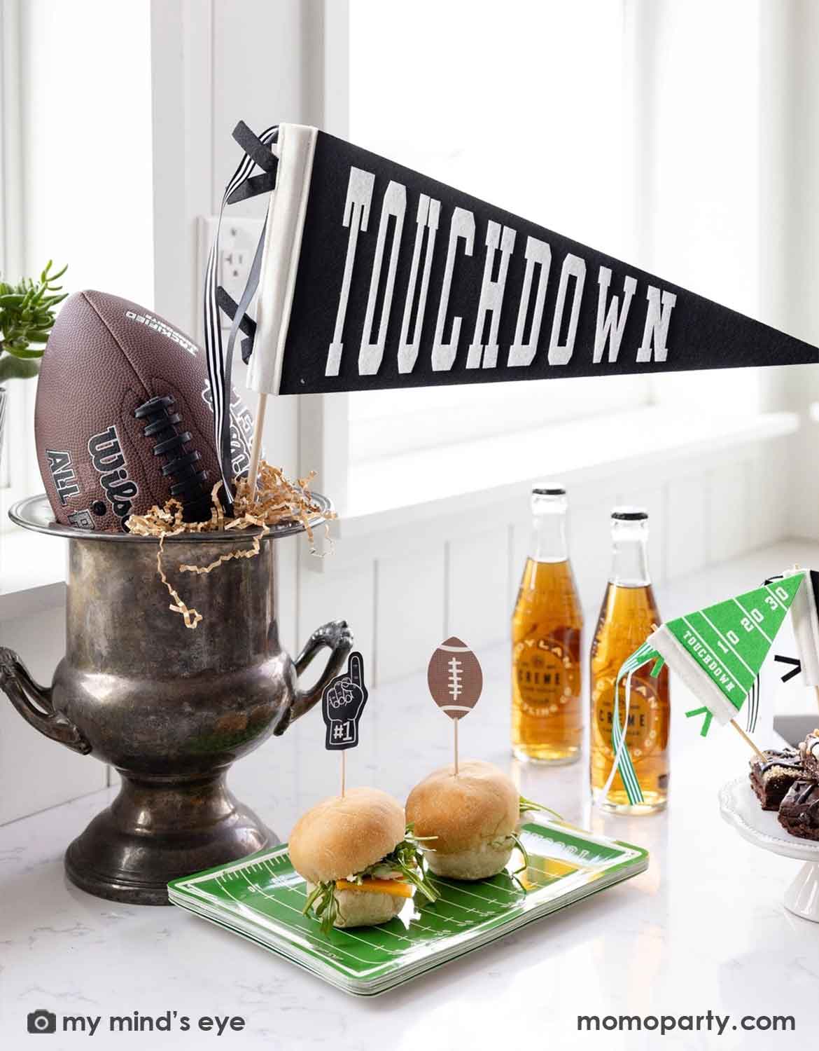 A football-themed party setup with Momo Party's black and white 'TOUCHDOWN' felt party pennant, a football in a silver bucket, two sliders on a green field-patterned plate filled with sliders, and beer bottles on a white table.