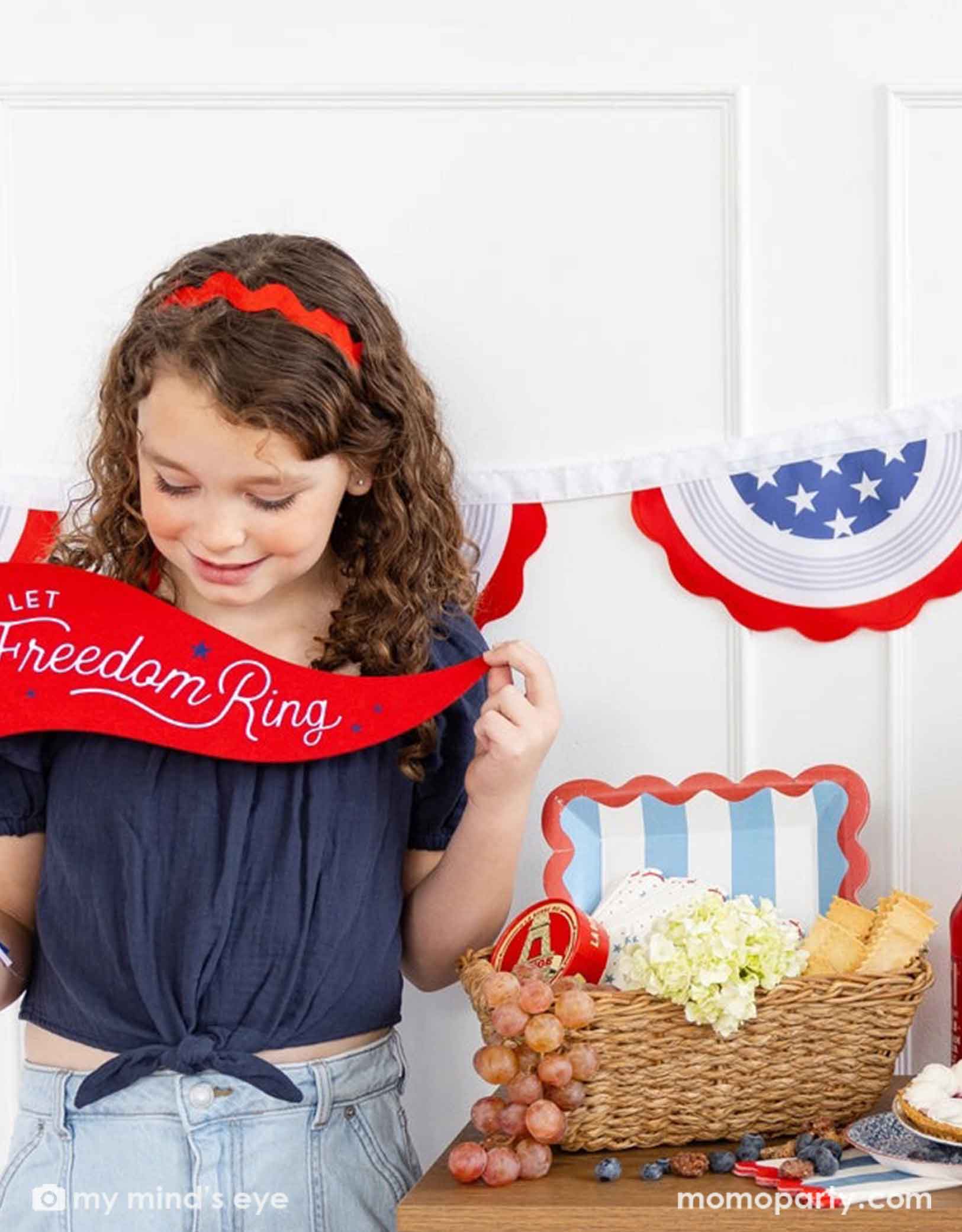 A young girl holding a red banner with 'Let Freedom Ring' text, standing in front of a patriotic-themed backdrop.
