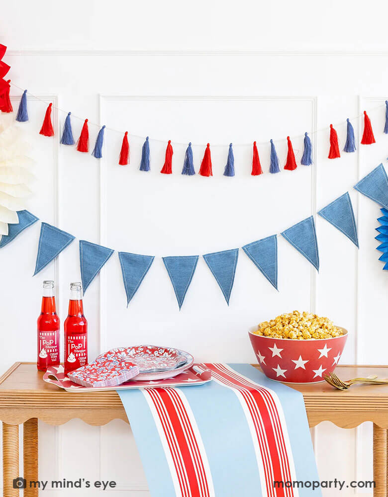 A wall decorated with Momo Party's patriotic party decorations and tableware by My Mind's Eye. Including a red, white and blue tassel banner, a blue flag banner with party paper fans. On the table there's the Hampton red and blue striped paper table runner, a red star bamboo tray with Hampton floral plates and napkins, next to two red soda bottles. On the right there's a big red star bamboo bowl with popcorn in it. Making this a great inspo for an elegant 4th of July home decor.