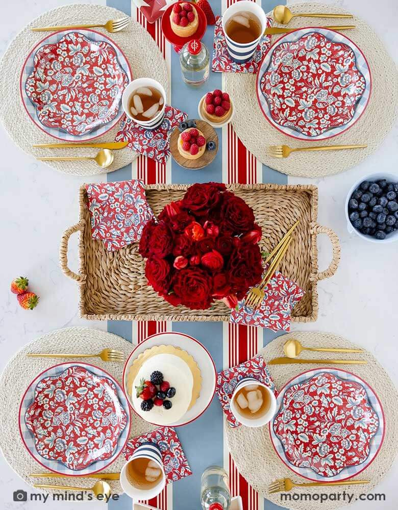 An elegant patriotic American themed party table features Momo Party's Hamptons Party Collection by My Mind's Eye including 10" Chambray Gingham Paper Plates, 10" Hampton Floral Wave Plates and napkins on white round placemats. In the middle of the table there are blue and red cakes, treats and drinks, along with a red floral bouquet as the centerpiece in the center on the Hampton striped paper runner, making this a perfect tablescape inspo for a 4th of July party or Memorial Day celebration.
