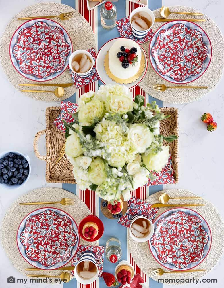 An elegant patriotic American themed party table features Momo Party's Hamptons Party Collection by My Mind's Eye including 10" Chambray Gingham Paper Plates, 10" Hampton Floral Wave Plates and napkins on white round placemats. In the middle of the table there are blue and red cakes, treats and drinks, along with a white floral bouquet as the centerpiece in the center on the Hampton striped paper runner, making this a perfect tablescape inspo for a 4th of July party or Memorial Day celebration.