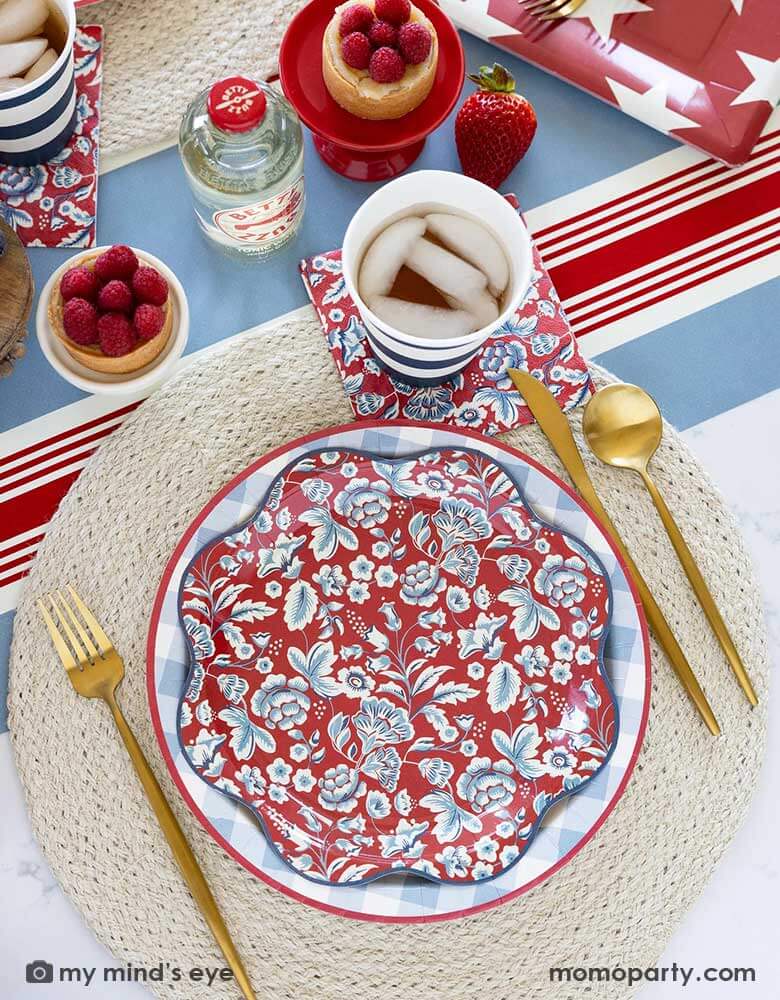 An elegant Fourth of July party table features Momo Party's Hamptons Collection by My Mind's Eye including 10" Chambray Gingham Paper Plate, Hampton Floral Wave Plate and napkin on a round white placemat. In the middle of the table there are blue and red treats and drinks on the Hampton striped paper runner, making this a perfect party inspo for a 4th of July party or Memorial Day celebration.
