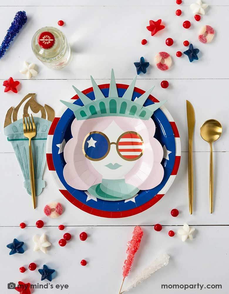A modern and fun placeset featuring Momo Party's Lady Liberty Party Collection by My Mind's Eye including the 10" red striped round plate, 9" blue star round plate, lady liberty shaped plate, torch shaped dinner napkins and. Around them are various candies and treats in patriotic themed in red white and blue. A perfect party inspo for a modern Fourth of July celebration with friends and family.