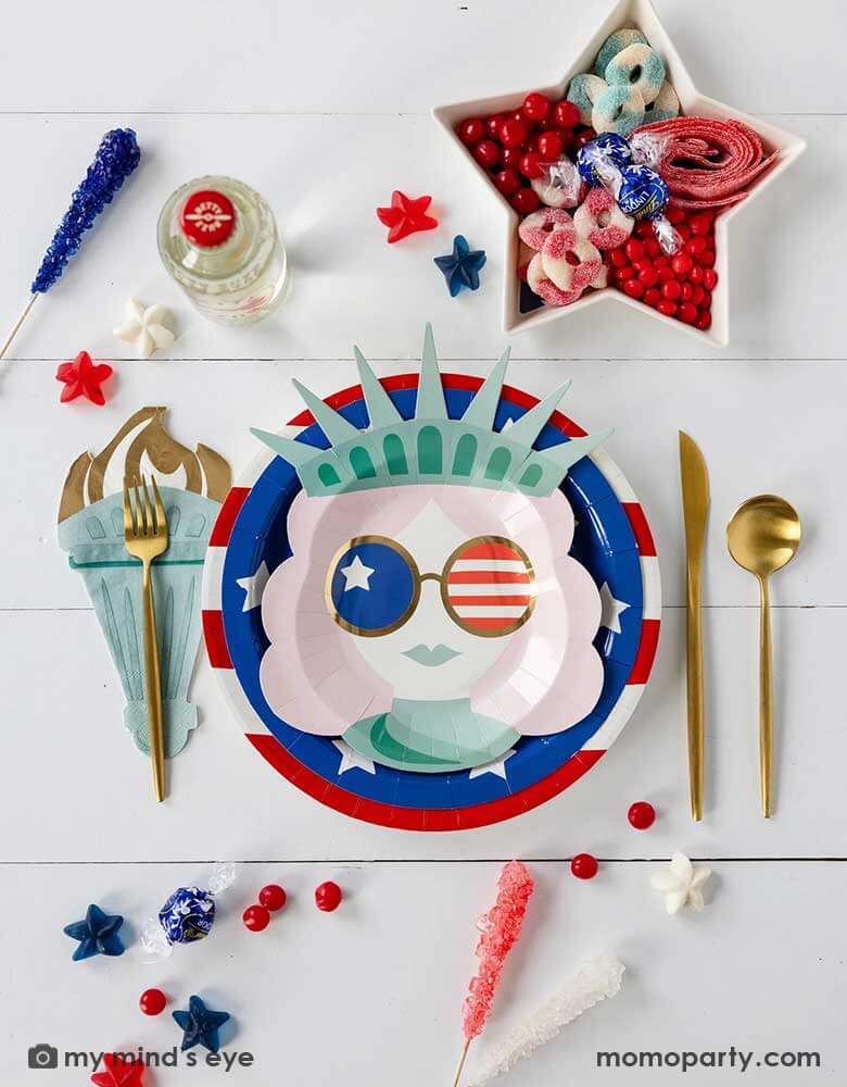 A modern and fun placeset featuring Momo Party's Lady Liberty Party Collection by My Mind's Eye including the 10" red striped round plate, 9" blue star round plate, lady liberty shaped plate, torch shaped dinner napkins and. Around them are various candies and treats in patriotic themed in red white and blue. A perfect party inspo for a modern Fourth of July celebration with friends and family.