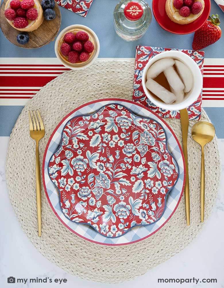 An elegant Fourth of July party table features Momo Party's Hamptons Collection by My Mind's Eye including 10" Chambray Gingham Paper Plate, Hampton Floral Wave Plate and napkin on a round white placemat. In the middle of the table there are blue and red treats and drinks on the Hampton striped paper runner, making this a perfect party inspo for a 4th of July party or Memorial Day celebration.