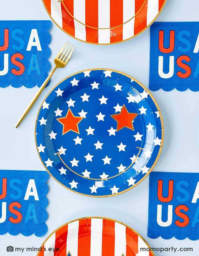 A light blue table features Momo Party's Star & Stripe Smiley Paper Plates by My Mind's Eye. These plates come in a set of 8 in 2 designs, one with red stripes the other with blue stars. With a big smiley face and star eyes on them. Pair with 5" USA small blue napkins, they are perfect for a retro fun 4th of July party this summer!