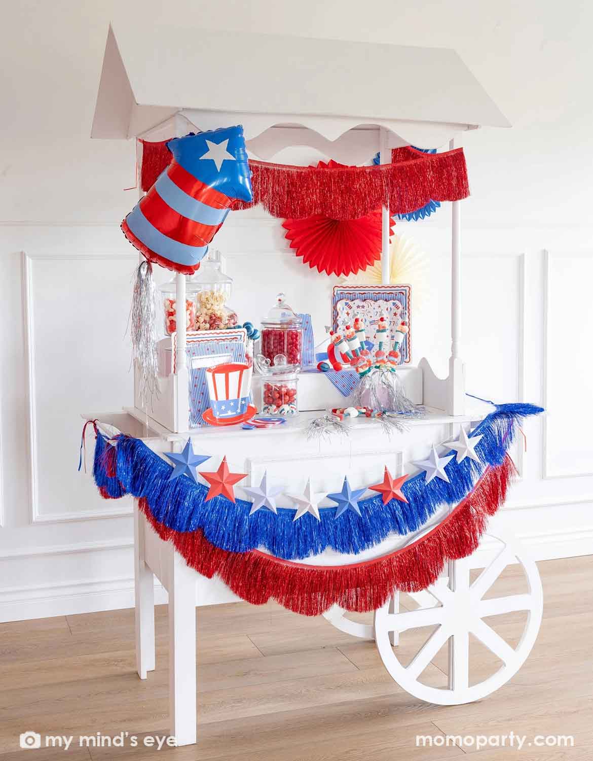 A patriotic themed decorated dessert cart with various American themed decors and tablewares in red, white, and blue from Momo Party, including paper fans, fringe bunting garlands, star party banners, a firecracker shaped foil balloon, plates, napkins and cups. A perfect inspiration for Fourth of July, Memorial Day, or summer celebrations with children.