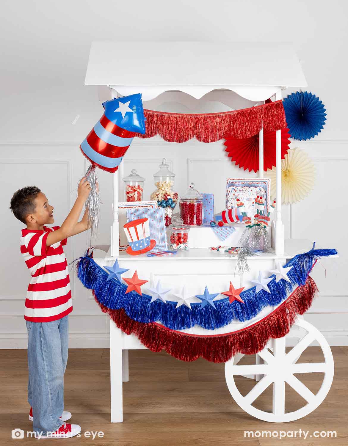 A patriotic themed decorated dessert cart with various American themed decors and tablewares in red, white, and blue from Momo Party, including paper fans, fringe bunting garlands, star party banners, a firecracker shaped foil balloon, plates, napkins and cups, along a boy in red, white striped outfits standing next to it playing with the balloon. A perfect inspiration for Fourth of July, Memorial Day, or summer celebrations with children.