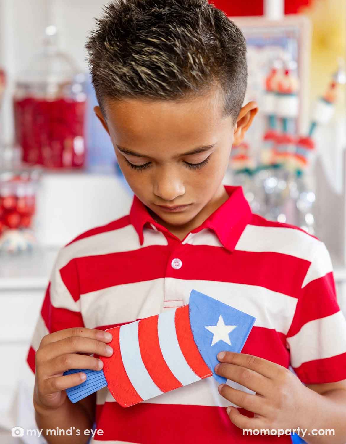 A boy in red striped polo shirt holding Momo Party's firecracker shaped napkin with American flag design by My Mind's Eye at a Fourth of July party decorated with festive patriotic themed decors in the back.