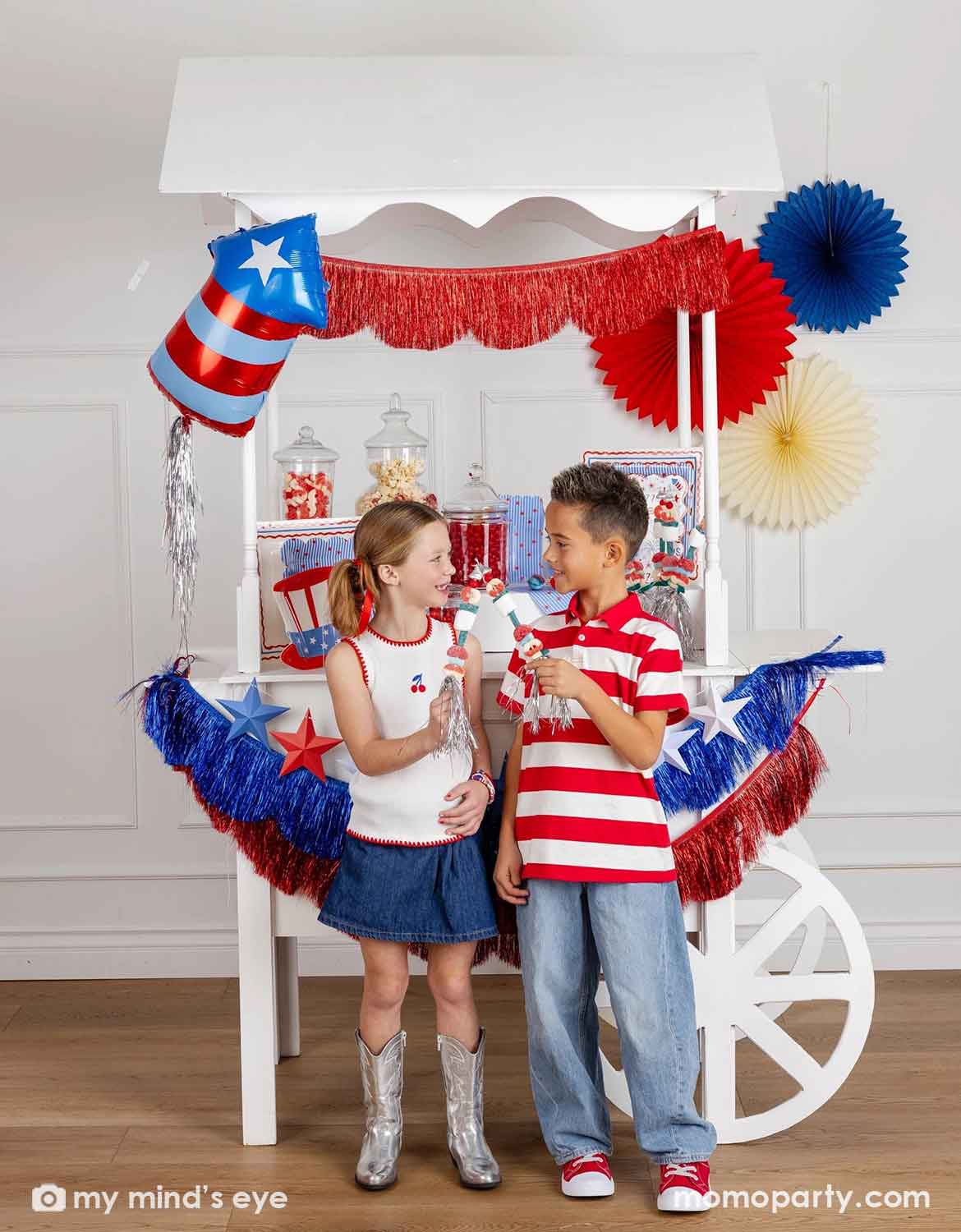 Two children in patriotic outfits standing in front of a Fourth of July themed decorated dessert cart with various American themed decors and tablewares in red, white, and blue from Momo Party, including paper fans, fringe bunting garlands, star party banners, a firecracker shaped foil balloon, plates, napkins and cups, along with a girl in blue shirt and skirt standing next to it. A perfect inspiration for Fourth of July, Memorial Day, or summer celebrations with kids.