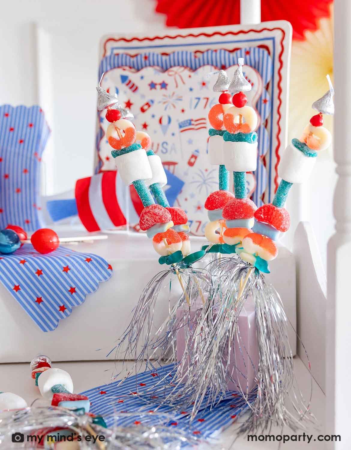 An USA themed party table setting featuring decorative candy kabobs in red, white and blue candies and marshmallow. In the back there are various American themed festive tableware from Momo Party along with some candy jars, a perfect setting for a kid's friendly Fourth of July party, Memorial Day celebration or a summer gathering.