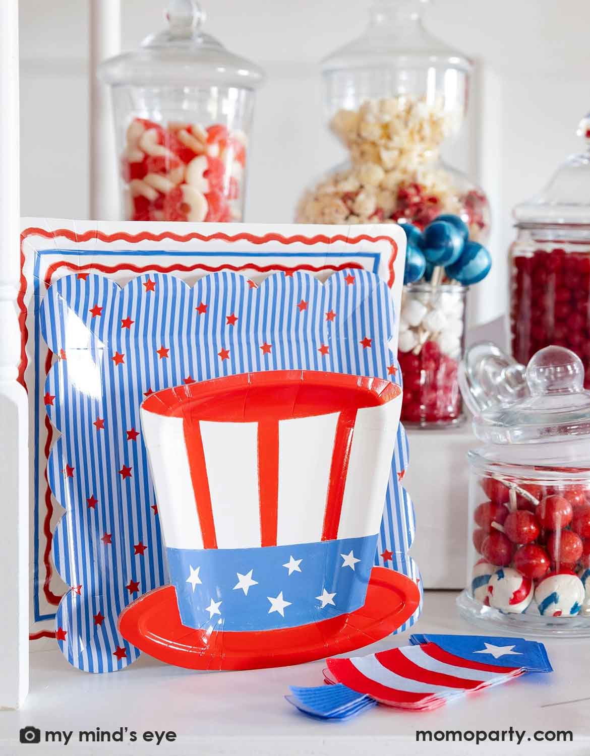 An USA themed party table setting featuring multiple American themed tablewares from Momo Party including stars and stripes plates, uncle Sam hat shaped plate and firecracker shaped napkins, along with candy jars filled with patriotic themed treats, it makes a perfect setting for a kid's friendly Fourth of July party, Memorial Day celebration or a summer gathering.