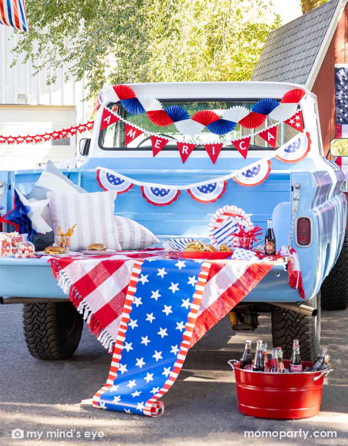 A light blue decorated Ford truck bed with multiple USA, American Fourth of July-themed decorations from Momo Party including bunting garlands, red flag banners and American flag buntings, along with various USA themed tablewares, coke bottles and snacks, a perfect set up for a festive Memorial Day party or Fourth of July celebration.