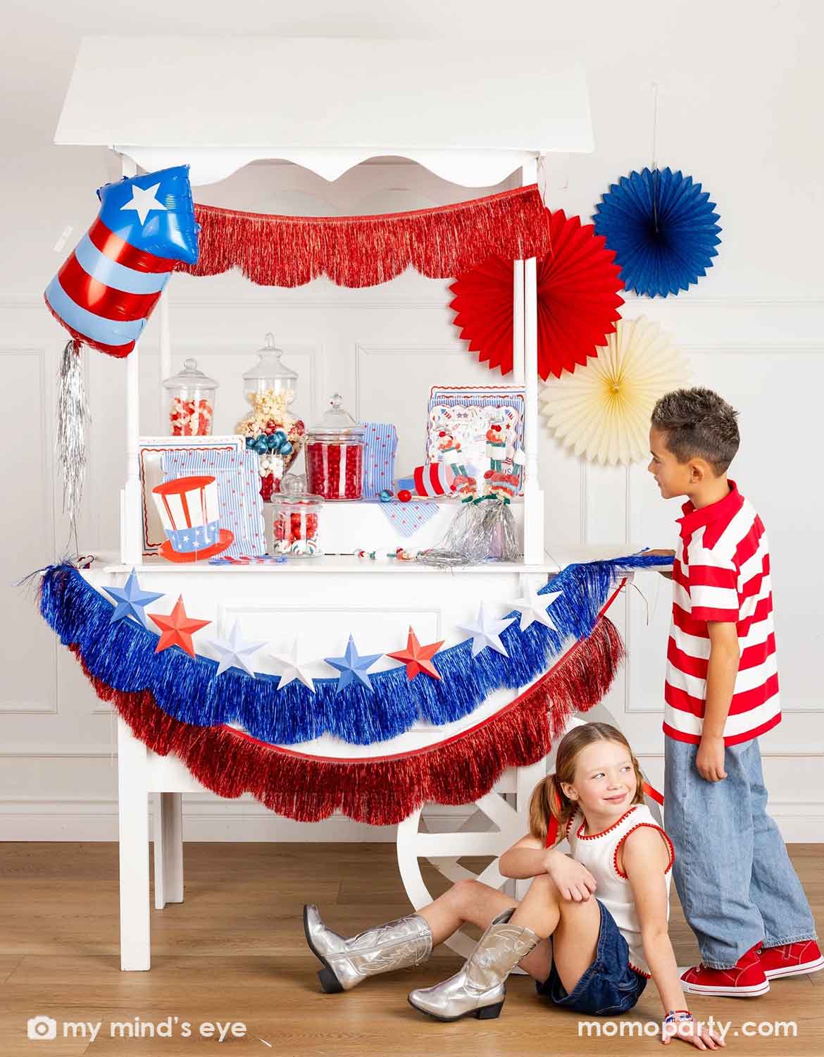 A patriotic themed decorated dessert cart with various American themed decors and tablewares in red, white, and blue from Momo Party, including paper fans, fringe bunting garlands, star party banners, a firecracker shaped foil balloon, plates, napkins and cups, along with two kids in red, white and blue outfits standing next to it. A perfect inspiration for Fourth of July, Memorial Day, or summer celebrations with children.