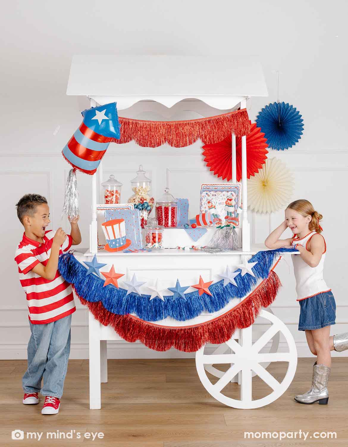 A patriotic themed decorated dessert cart with various American themed decors and tablewares in red, white, and blue from Momo Party, including paper fans, fringe bunting garlands, star party banners, a firecracker shaped foil balloon, plates, napkins and cups, along with two kids in red, white and blue outfits standing next to it. A perfect inspiration for Fourth of July, Memorial Day, or summer celebrations with children.