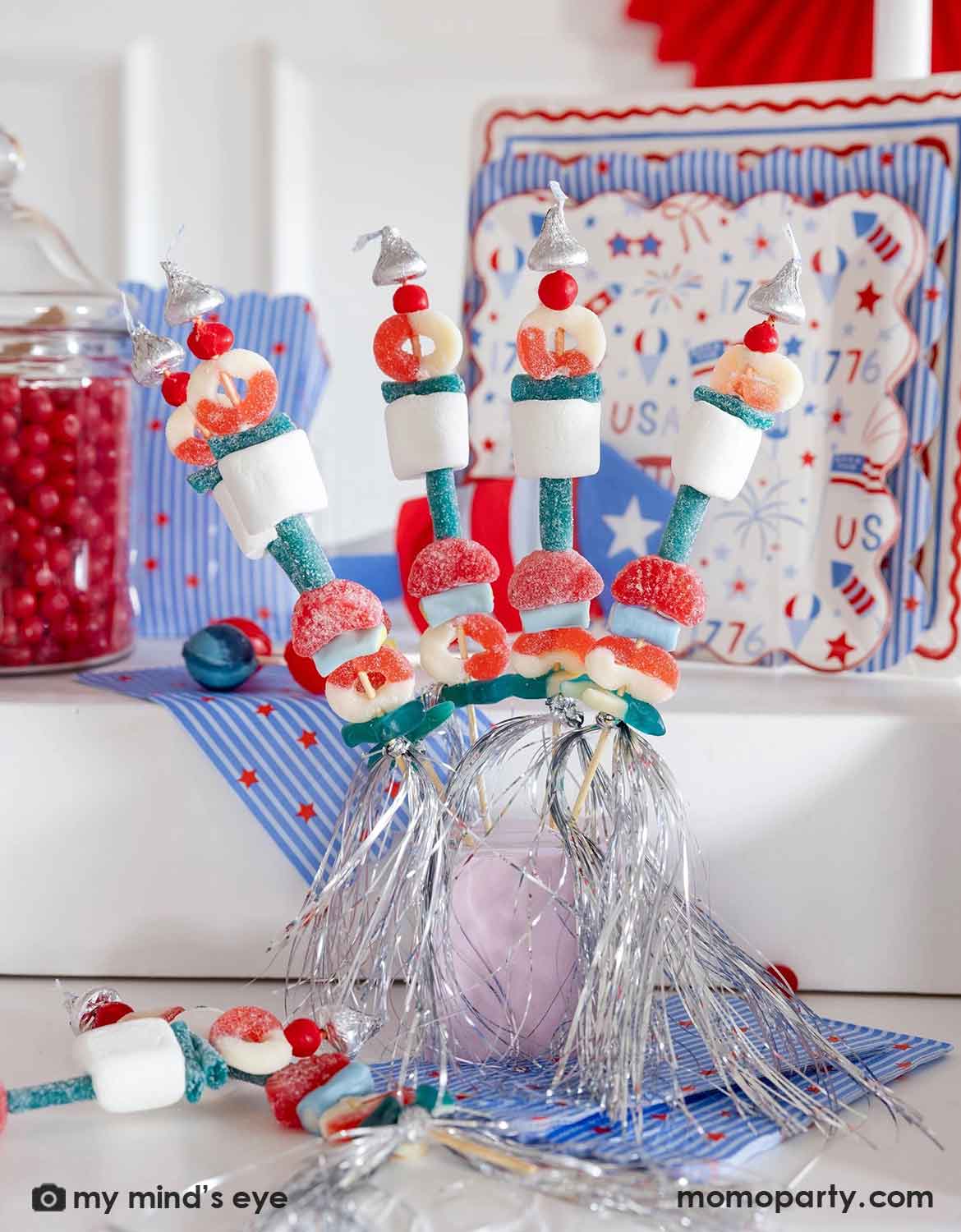 An USA themed party table setting featuring decorative candy kabobs in red, white and blue candies and marshmallow. In the back there are various American themed festive tableware from Momo Party along with some candy jars, a perfect setting for a kid's friendly Fourth of July party, Memorial Day celebration or a summer gathering.