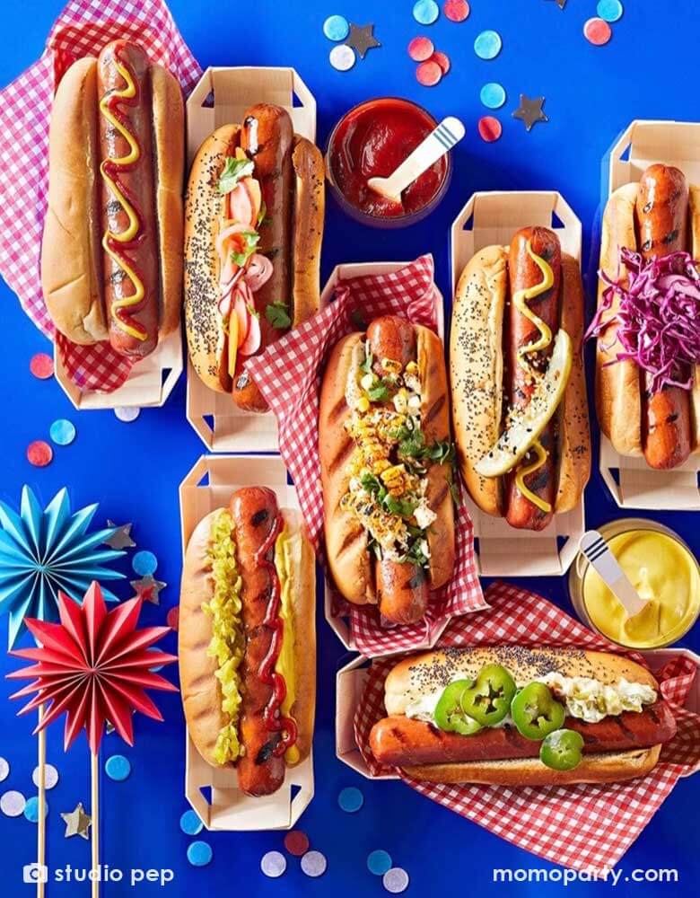 A table filled with different flavor of hotdogs with various condiment in hotdog paper containers adorned with red checkered paper, on the blue party table is Momo Party's patriotic confetti in red, white and blue spread out and some mini paper fan toppers in blue and red, making this a perfect inspo for a festive fourth of July party table and food ideas.