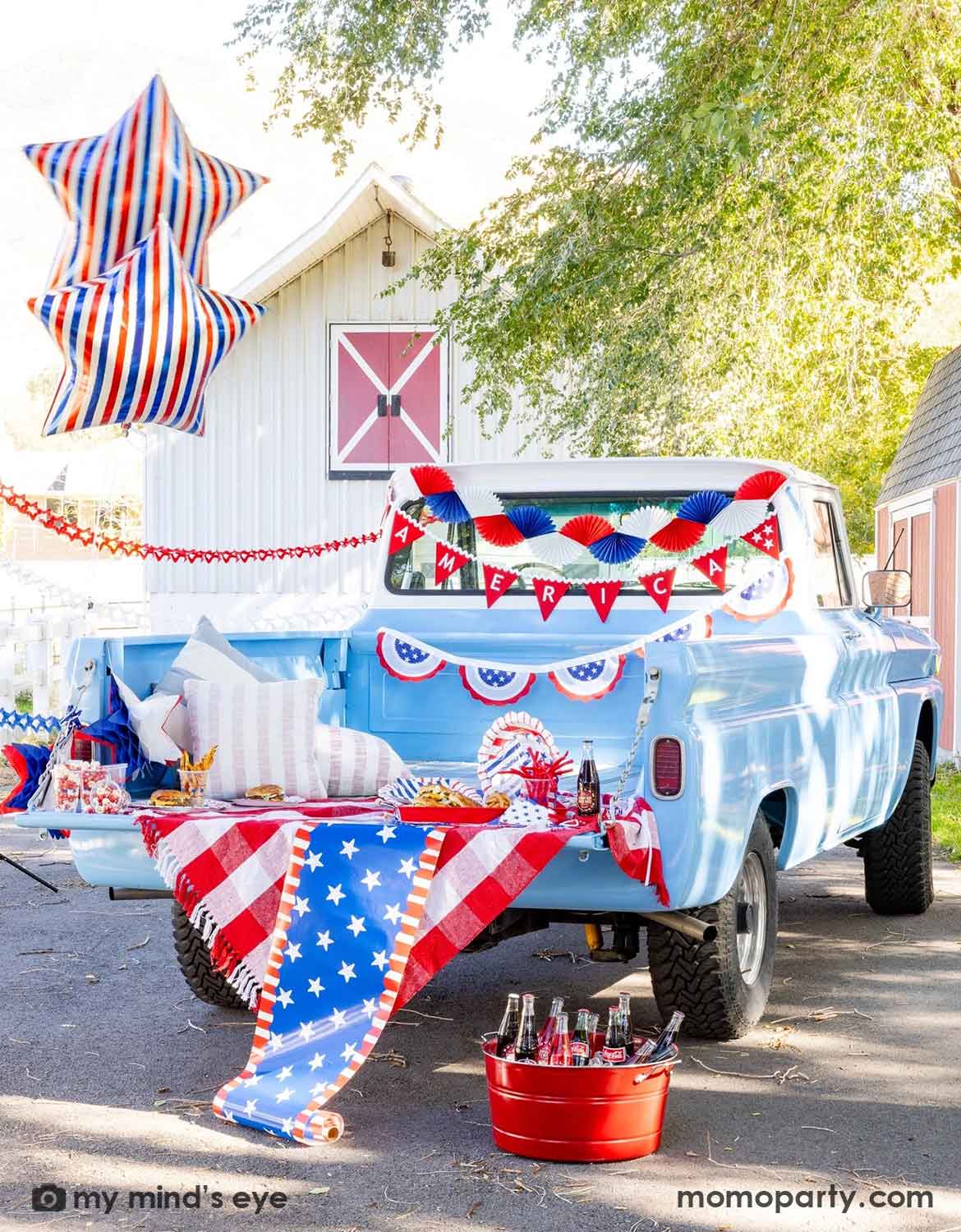 A light blue decorated Ford truck bed in a driveway with multiple USA, American Fourth of July-themed decorations and festive foil star shaped balloons from Momo Party, including bunting garlands, red flag banners and American flag buntings, along with various USA themed tablewares and table runner with coke bottles and snacks on it, a perfect set up for a festive Memorial Day party or Fourth of July celebration.