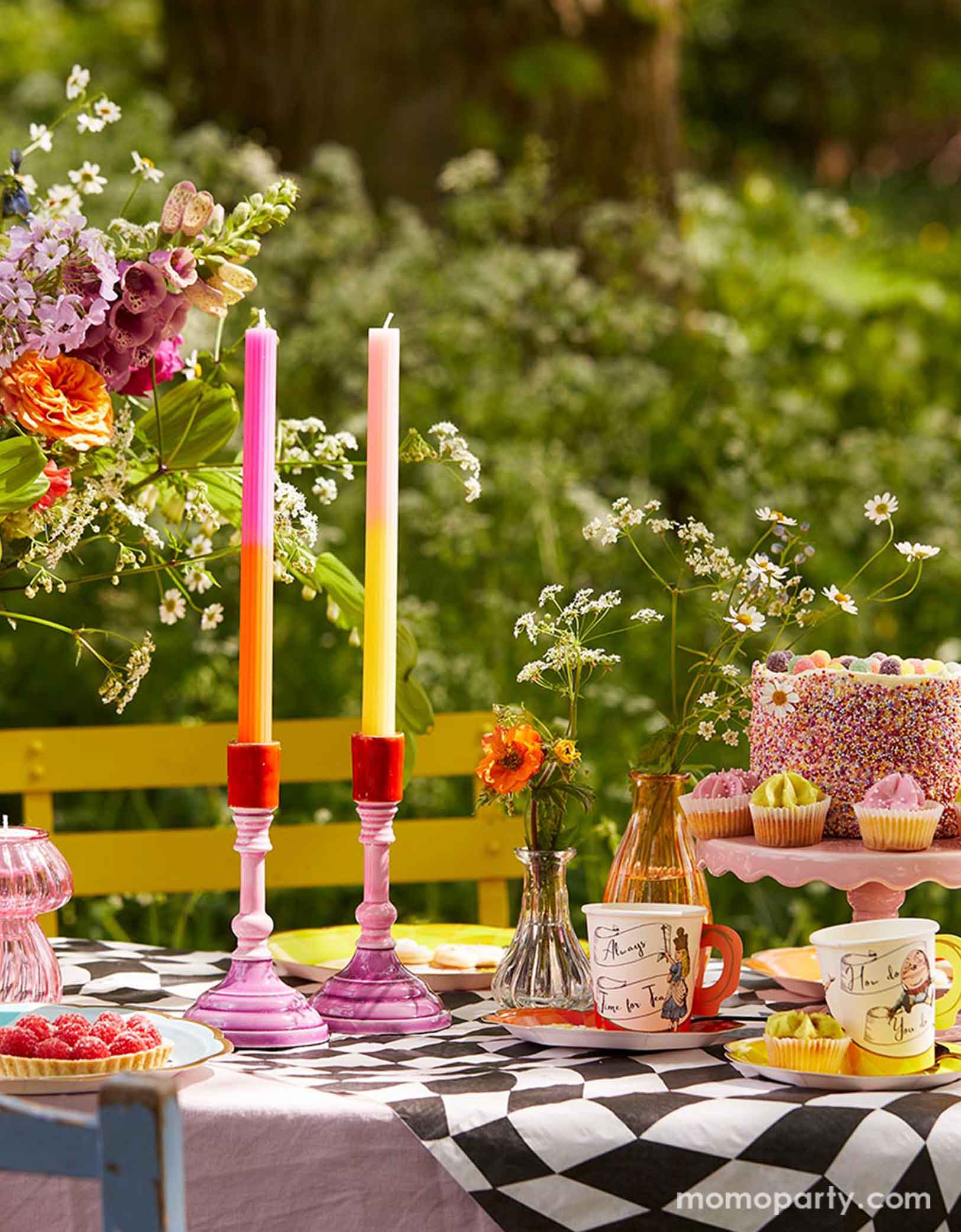 An Alice Wonderland themed garden tea party table set up featuring whimsical party supplies including plates, party cups and napkins on the black and white check table cover from Momo Party. With some flowers in glass vases, a birthday cake, some cupcakes, desserts, long candles on the table, a magical journey is about to start!