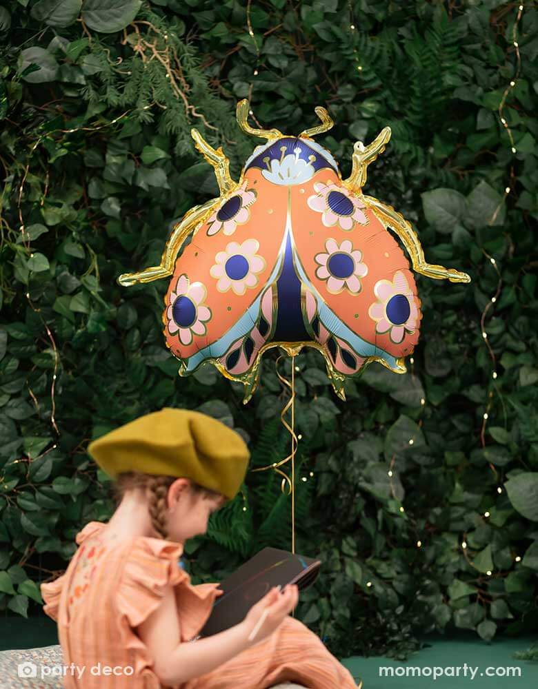 A little girl drawing on her sketchbook in the backyard with a wall of greenery, next to her is Momo Party's 34 x 30 inches ladybird shaped foil balloon by Party Deco. With coral color with small flower pattern with gold legs on it.