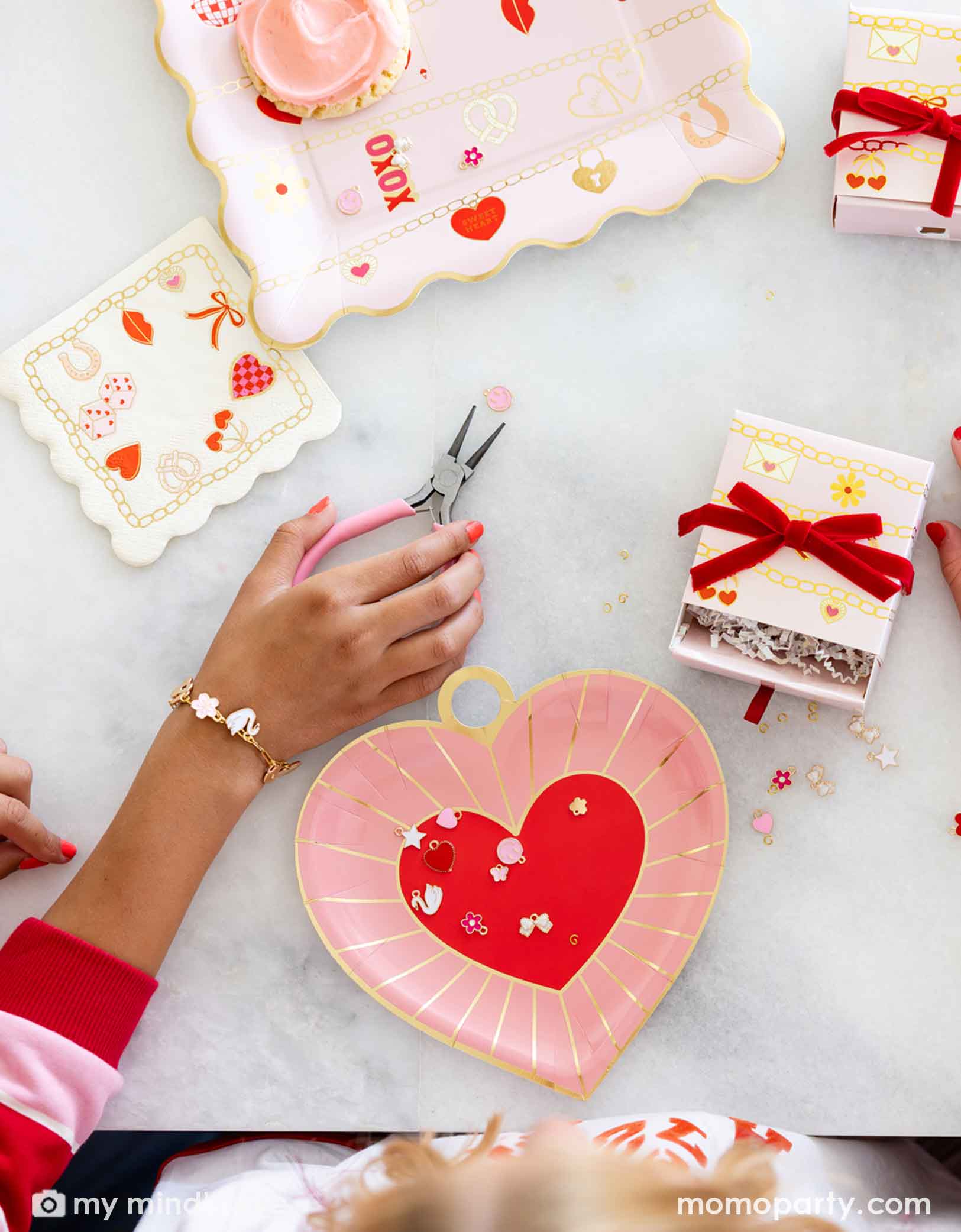 Children's hands with pink nail polish working on their charm bracelets with pink pliers, surrounded by small charms, gift boxes and Momo Party's Valentine's Day charm matching party tablewares including heart shaped plates and napkins on the table.